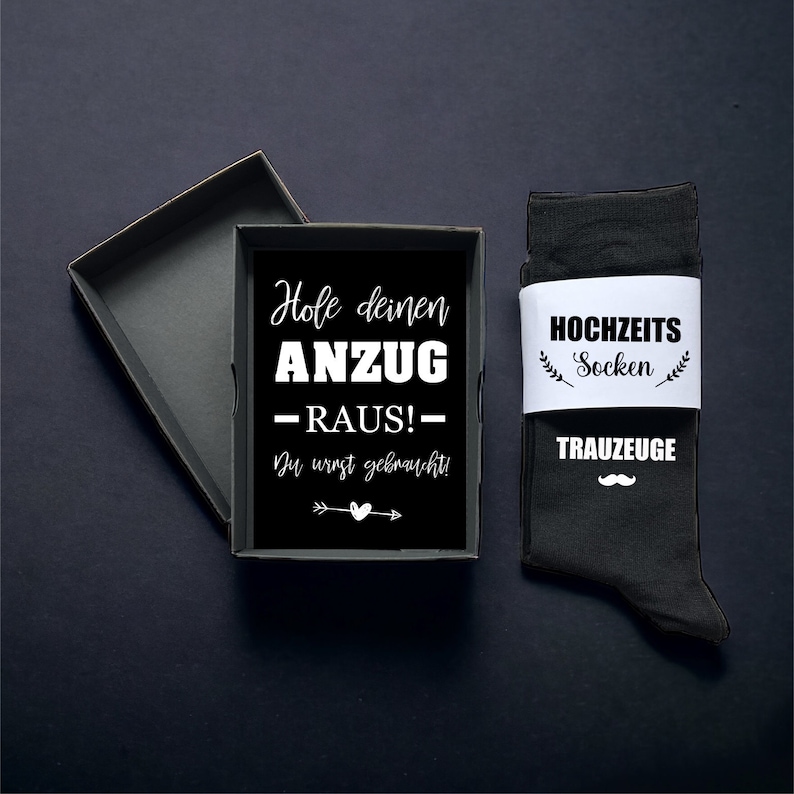 May include: A black gift box with a black interior, next to a pair of black socks with a white band. The box contains a black card with white text that reads "Hole deinen ANZUG - RAUS!" and "Du wirst gebraucht!" The socks say "HOCHZEITS Socken" and "TRAUZEUGE".