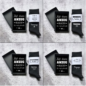 May include: Four black gift boxes, each containing a pair of black socks and a card. The cards read "Hole deinen ANZUG - RAUS!" and "Die wirst gebraucht!". The socks have text like "Trauzeuge".