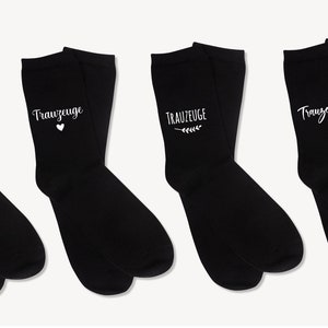 May include: Four pairs of black socks, each bearing the white text "TRAUZEUGE". The socks feature different embellishments: a moustache, a heart, a sprig, and a simple script. These socks are likely intended for a wedding.