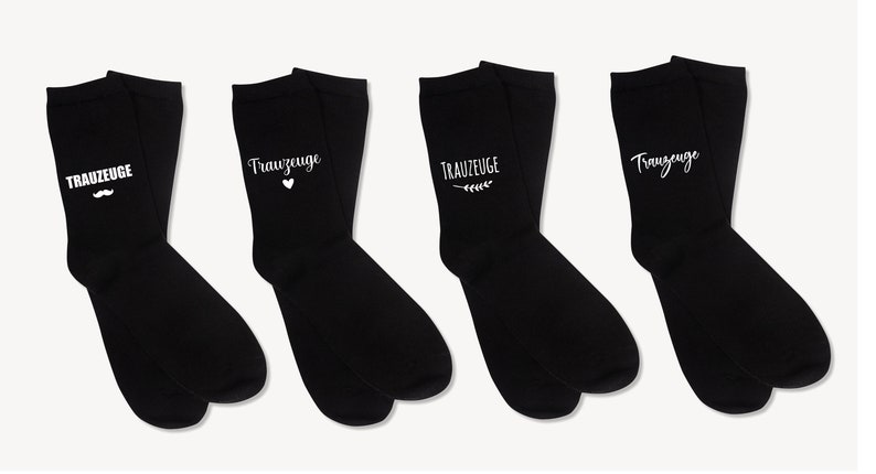 May include: Four pairs of black socks, each bearing the white text "TRAUZEUGE". The socks feature different embellishments: a moustache, a heart, a sprig, and a simple script. These socks are likely intended for a wedding.