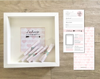 May include: A white frame with a pink and white card inside that says "Zeitreise" and "Bitte am 12.08.2027 öffnen". There are five rolled up pieces of paper inside the frame. There are two white cards with black text and a pink card with pink hearts on it.