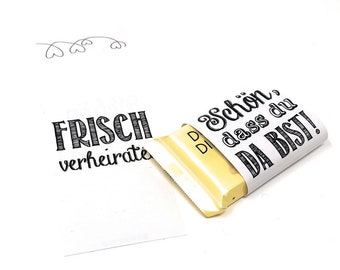 May include: A white chocolate bar wrapped in white paper with black text that reads "Schon, dass du DA BIST!"  The wrapper is partially peeled back to reveal the chocolate bar. A separate white card with black text that reads "FRISCH verheiratet" is also shown.