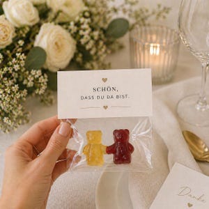 May include: A clear plastic bag containing one yellow and one red gummy bear holding hands. The bag is sealed with a white paper topper featuring two small hearts and the German text: SCH&Ouml;N, DASS DU DA BIST. The design is simple and minimalist.