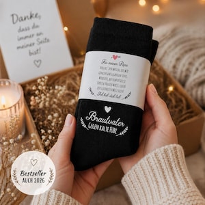 May include: Black socks with white text, held by a person. The socks have text that reads "Brautvater Gegen Kalte Füße" and a message for "Papa". A gift box and a candle are in the background. The image also includes the text "Bestseller AUCH 2026".