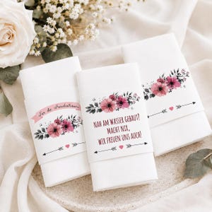 May include: Three white folded napkins with floral bands. The bands feature pink and purple flowers, green leaves, arrows, and hearts. Text on the bands reads "Far die Freudentränen" and "NAH AM WASSER GEBAUT? MACHT NIX, WIR FREUEN UNS AUCH!"