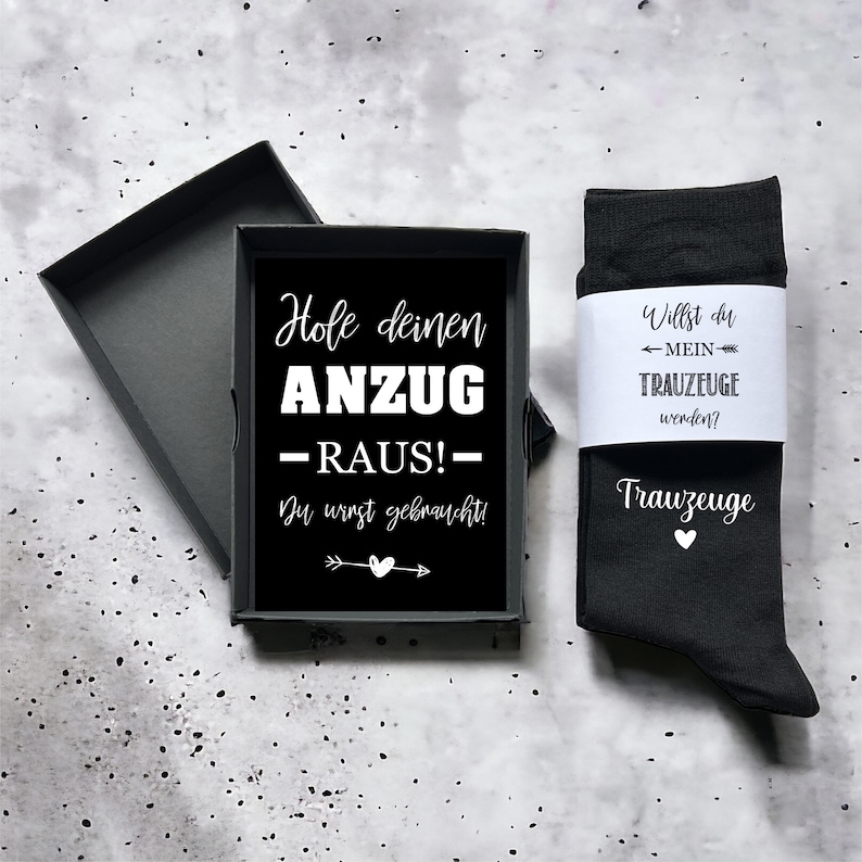 May include: A black gift box containing a black card with white text in German and a pair of black socks. The socks have a white label with German text and the word "Trauzeuge" printed on them.