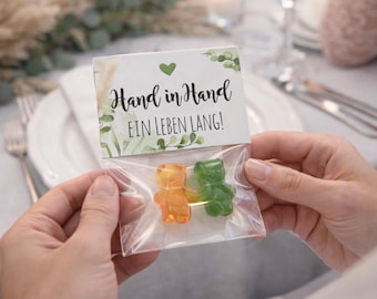May include: A clear plastic bag containing two gummy bear candies, one orange and one green. The bag is attached to a white card with the text "Hand in Hand EIN LEBEN LANG!" and a green heart and leaf design. The image is taken indoors.