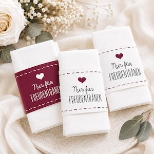 May include: Three white folded napkins with a burgundy band featuring a heart and the text "Nur für Freudentränen". The napkins are arranged on a white surface, with flowers in the background.