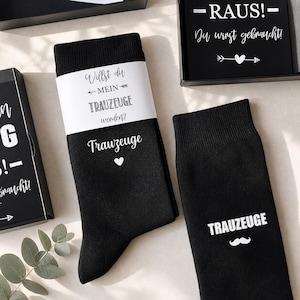 May include: Black socks with white text and a heart, and a mustache. One sock has the word "Trauzeuge" and a heart. The other sock has the word "Trauzeuge" and a mustache. A white band around the socks reads "Willst du MEIN TRAUZEUGE werden?"