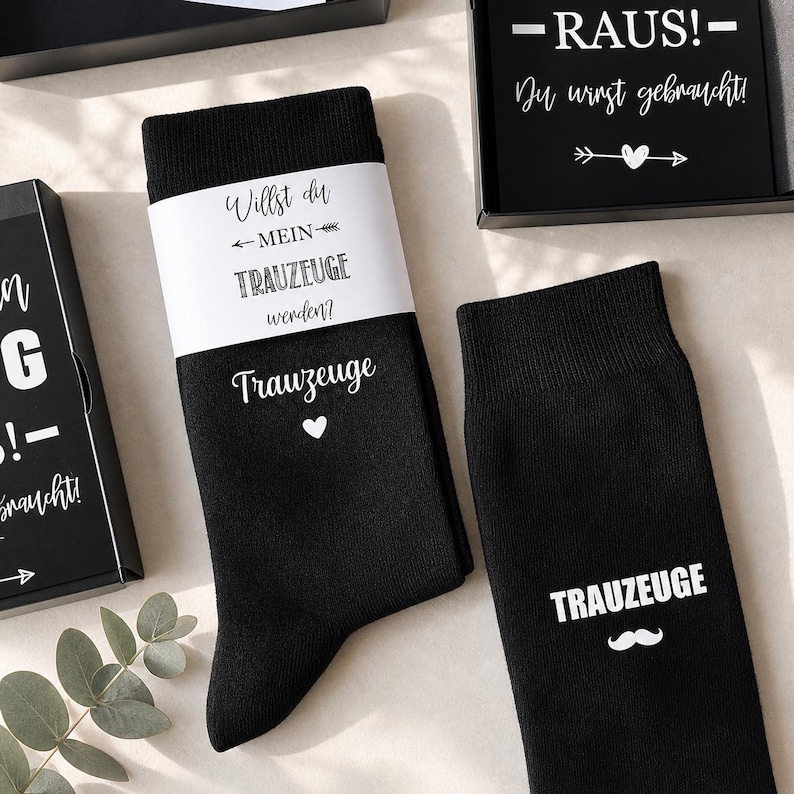May include: Black socks with white text and a heart, and a mustache. One sock has the word "Trauzeuge" and a heart. The other sock has the word "Trauzeuge" and a mustache. A white band around the socks reads "Willst du MEIN TRAUZEUGE werden?"