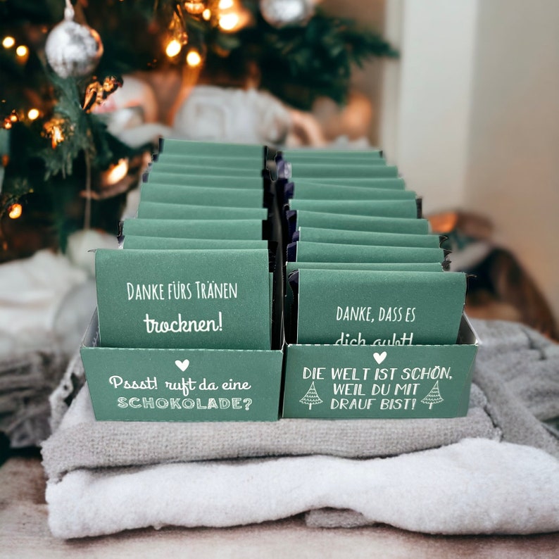 May include: A collection of small green boxes with white text. The boxes have messages like "Danke f&uuml;rs Tr&auml;nen trocknen!" and "Pssst! ruft da eine Schokolade?"  The boxes are arranged in a rectangular shape and are sitting on a white and gray surface.