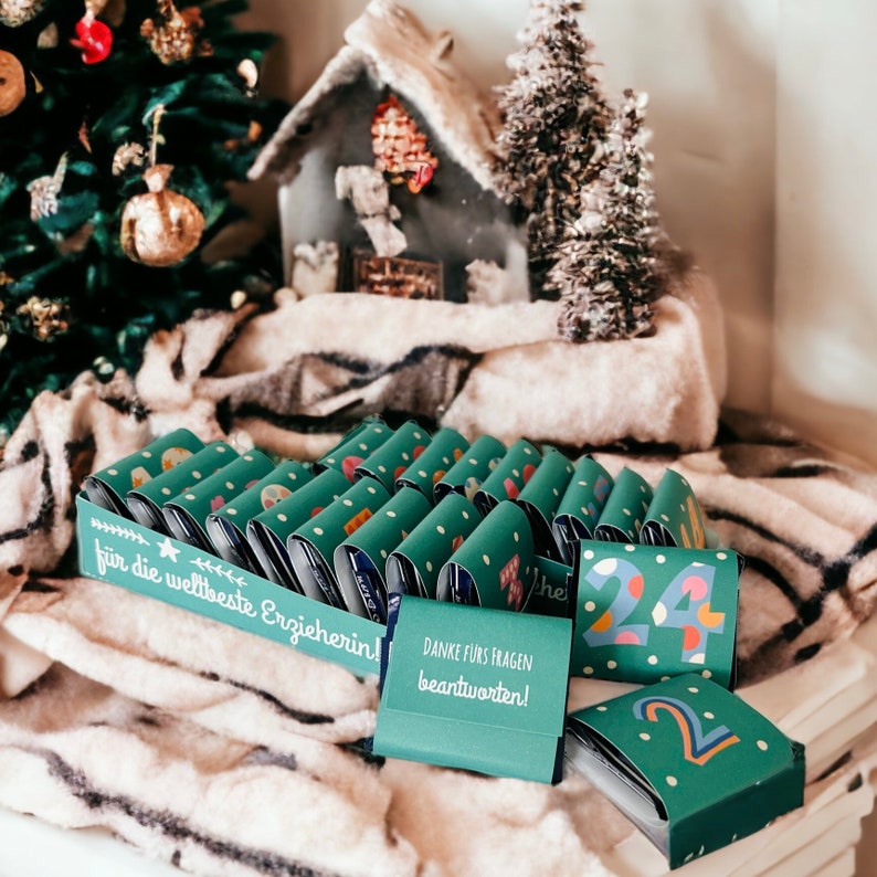 May include: A green and blue advent calendar with 24 small boxes. The boxes are decorated with white polka dots and have different phrases written on them, including "Danke f&uuml;rs Fragen beantworten!" and "f&uuml;r die weltbeste Erzieherin!"