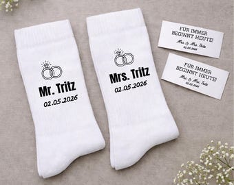 May include: White wedding socks with the text "Mr. Tritz" and "Mrs. Tritz" along with the wedding date 02.05.2026. Each sock features a ring graphic. Two small cards with German text are also present.