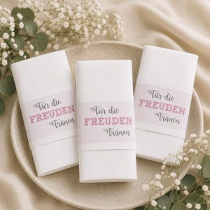 May include: Three folded white napkins with a pink and gray band that reads "Für die Freuden Tränen". The napkins are arranged on a light beige plate, surrounded by white flowers and greenery.