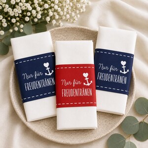 May include: Three white folded napkins with decorative bands in navy blue and red. The bands feature the text "Nur für Freudentränen" with a heart and anchor design. The napkins are arranged on a beige plate.