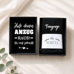 May include: Black gift box with white text that reads "Hole deinen ANZUG -RAUS!- Du wirst gebraucht!" and a small arrow with a heart. Next to it is a black sock with the word "Trauzeuge" and the name "Marcel".