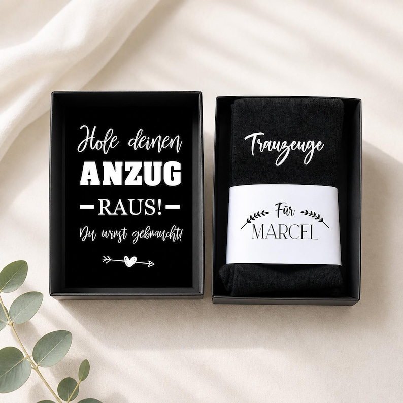 May include: Black gift box with white text that reads "Hole deinen ANZUG -RAUS!- Du wirst gebraucht!" and a small arrow with a heart. Next to it is a black sock with the word "Trauzeuge" and the name "Marcel".