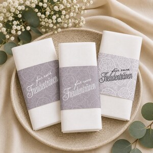 May include: Three white paper napkins with a gray floral band and the text "für eure Freudentränen" in elegant script. The napkins are arranged on a beige plate, with greenery in the background.
