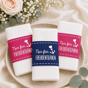May include: Three white folded napkins with decorative bands. Two bands are hot pink, one is navy blue. Each band has the text "Nur für Freudentränen", a heart, and an anchor. The napkins are on a beige plate.