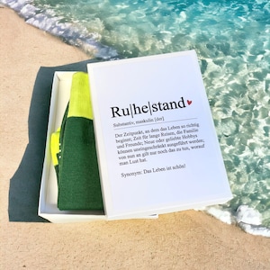 May include: A white box containing a pair of green and yellow socks. The box features a white card with the word "Ruhestand" in bold black lettering and a definition in German.