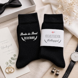 May include: A pair of black socks with white text, one reading "Bruder der Braut Bester Bruder" and the other "HOCHZEITSSOCKEN Lieblingsgast". A wedding invitation, gold rings, and a watch are also visible.