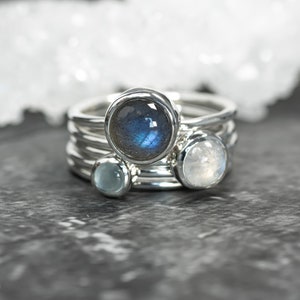 May include: A stack of four silver rings, each with a different gemstone. The top ring features a large, round, blue-green labradorite stone. Other rings have a light blue, and a white stone. The rings are stacked on a dark gray surface.