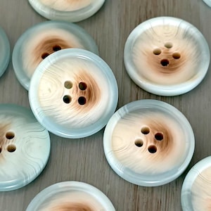 May include: A collection of twelve round, four-hole buttons. Each button features a light blue outer rim that transitions into a swirling, cream and brown wood-grain pattern toward the center, creating a distinct contrast across the set.