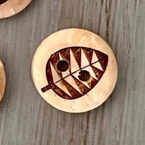 May include: Three round, light tan buttons, each featuring a dark brown, laser-engraved leaf design. Every button has two central holes positioned within the leaf pattern, showcasing a smooth, polished surface with natural variations in color.