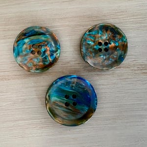 May include: Three round buttons with four holes each. The buttons have a marbled appearance with shades of teal, blue, and brown. The buttons are arranged on a light-colored wooden surface.
