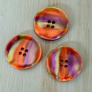 May include: Three round buttons featuring a swirling, marbled pattern of vibrant orange, deep purple, and soft yellow hues. Each button has a smooth, glossy finish and includes four centered holes for attachment, arranged in a square formation.