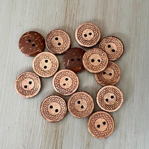May include: A collection of brown, round buttons with two holes each. Some buttons have a textured, woven-like pattern around the holes. The buttons vary in shade from light to dark brown, and are scattered on a light blue-gray surface.