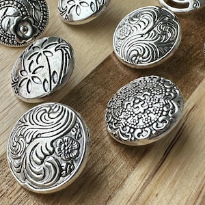 May include: Close-up of several silver-toned metal buttons with intricate floral and swirl designs. The buttons are oval and round, with detailed patterns. They are arranged on a light-colored wooden surface.