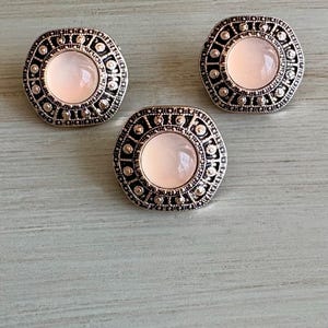 May include: Three silver-toned, square-shaped buttons with a central, light pink, circular stone. The buttons have an ornate, detailed border with small, clear accents. The buttons are arranged on a light blue-green surface.