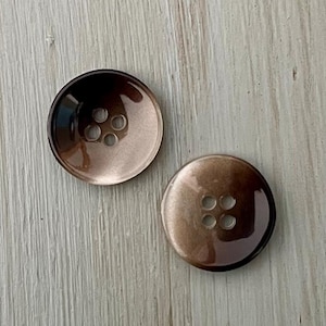 May include: Two brown plastic buttons with four holes each. The buttons have a gradient color from a darker brown to a lighter brown.
