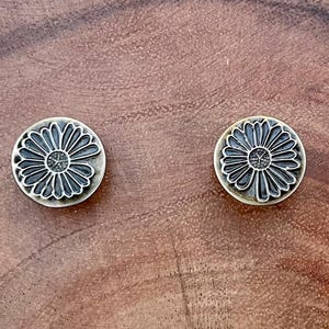May include: Two round, silver-toned metal earrings with a raised, detailed design of a daisy flower.
