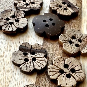 May include: A collection of flower-shaped buttons made from dark brown and light tan wood. The buttons have four holes for sewing and a detailed floral design carved into the surface. The buttons are arranged on a light brown wooden surface.