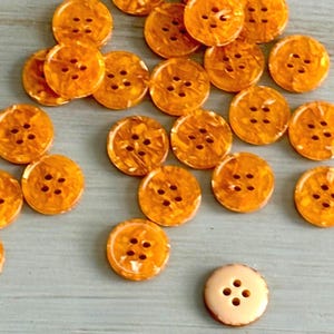 May include: A collection of round, four-hole buttons in a vibrant, translucent orange marbled pattern, scattered across the frame. Each button features a smooth, glossy finish with subtle variations in color depth, and one solid light beige button.
