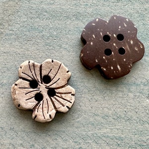 May include: Two flower-shaped buttons. One is a light gray color with black button holes and etched lines. The other button is dark brown with white speckles and black button holes. Both buttons have four holes.