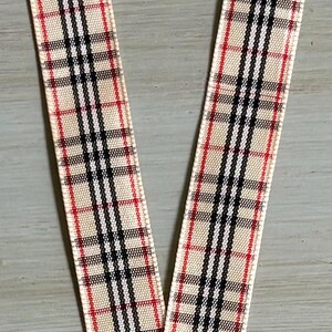 May include: Close-up of two beige ribbons with a classic plaid pattern in black, red, and white. The ribbons are likely for crafting, gift wrapping, or decorative purposes.