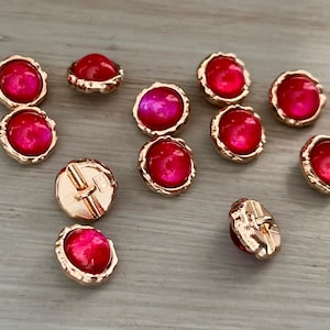 May include: A collection of decorative buttons with a gold-colored, textured rim and a vibrant, pinkish-red center. The buttons are scattered on a light-colored surface, showcasing their glossy finish and intricate design.