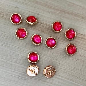 May include: A collection of decorative buttons with a gold-colored, wavy edge. The buttons have a vibrant, pink, glossy center. Two buttons show the back side with a cross-shaped design.