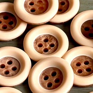 May include: A collection of round buttons featuring a smooth cream-colored outer rim and a recessed, textured brown center. Each button has four holes arranged in a square pattern in the middle, designed for sewing onto garments or fabric items.