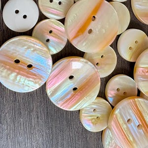 May include: A collection of round, two-hole buttons featuring a pearlescent finish with iridescent streaks of soft pink, orange, and cream. The buttons vary in size and display a smooth, glossy surface texture with subtle color variations across each piece.