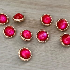 May include: A collection of nine round, decorative buttons with a vibrant pink center and a gold-colored, textured rim. The buttons are scattered on a light-colored wooden surface.