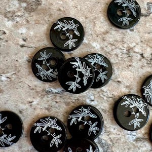 May include: A collection of round, black buttons, each featuring a white line-art illustration of bamboo stalks and leaves. The buttons are circular with two holes in the center for attachment, displaying a consistent botanical pattern.
