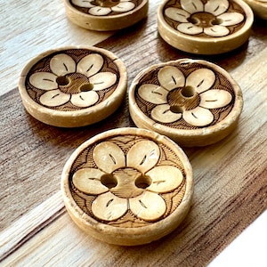 May include: A collection of light brown, wooden buttons with a floral design carved into the surface. Each button has a central hole and a series of petals around it. The buttons are arranged on a light brown and white wooden surface.