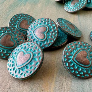 May include: A group of teal and copper heart-shaped buttons with a textured surface. The buttons are arranged in a pile on a wooden surface.
