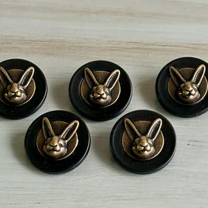 May include: Five black buttons with a bronze-colored rabbit head design. The rabbit is centered on a circular bronze background, set within a black button. These buttons are suitable for sewing or craft projects.