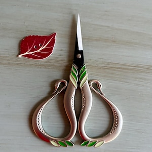 May include: Decorative embroidery scissors with swan-shaped handles and a red leaf-shaped button. The scissors have a black blade and are accented with green and cream-colored enamel. The handles are a metallic rose gold color.