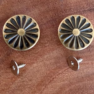 May include: Two gold-toned metal buttons with a daisy-like design and a black center. Each button comes with a silver screw.